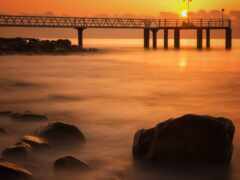 закат над пирсом с лодкой вдалеке и скалами на переднем плане