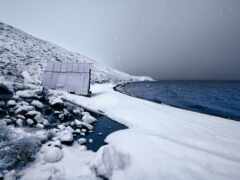 снежная сцена маленькой хижины на скалистом берегу с водоемом