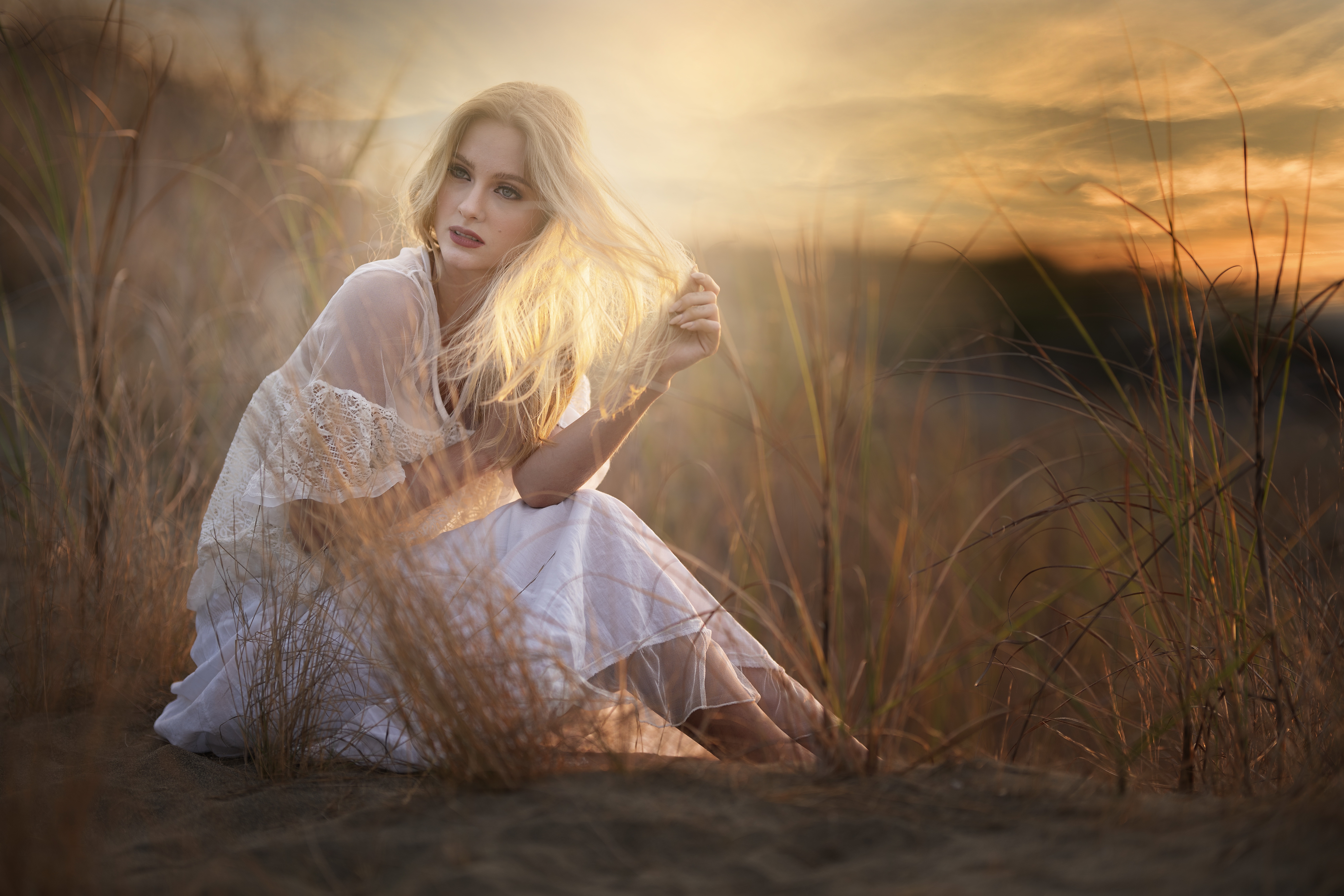 блондинка сидит на поле высокой травы с закатом на заднем плане