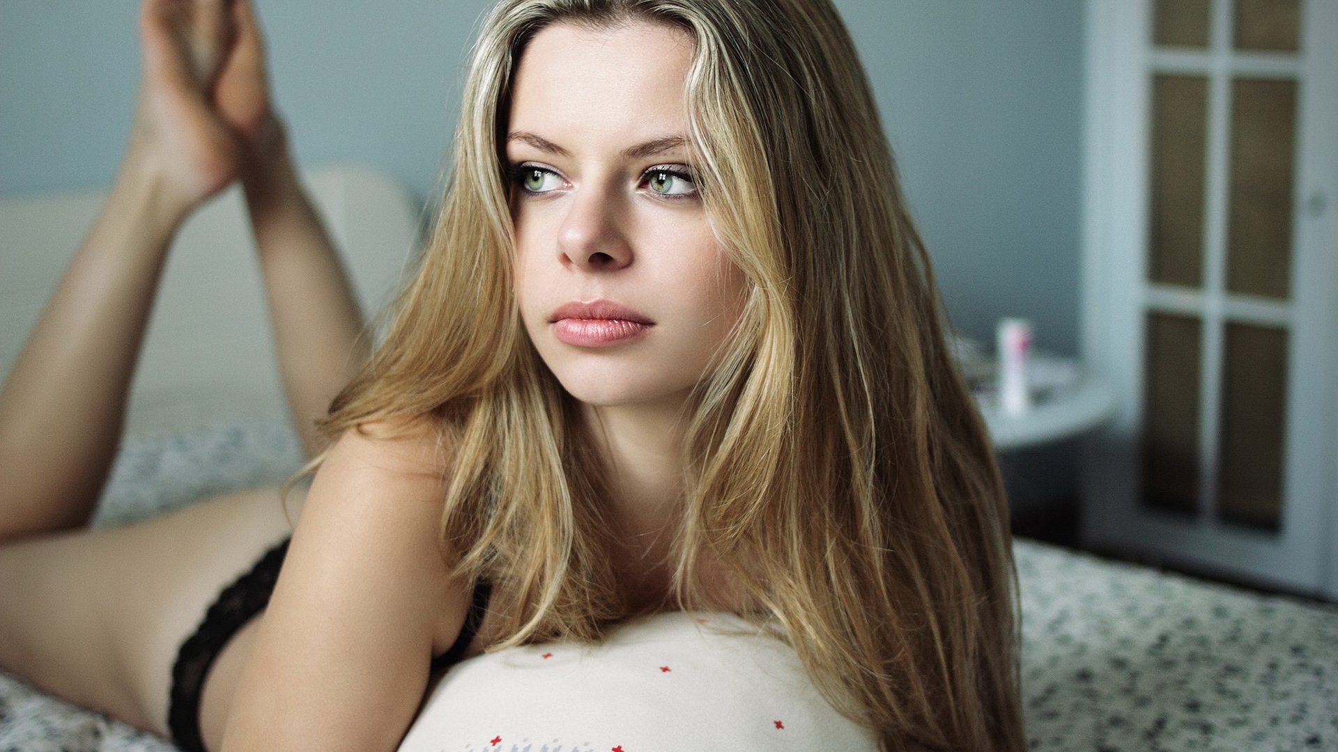 блондинка лежит на кровати со скрещенными ногами и смотрит в камеру