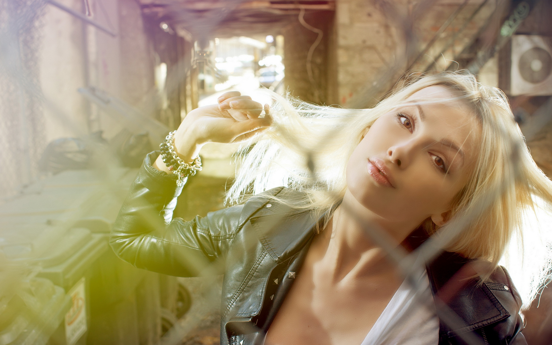 блондинка в кожаной куртке прислонилась к забору с выдувающимися на ветру волосами